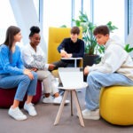 Vier junge Menschen sitzen in einem modernen, hellen Raum mit großen Fenstern und unterhalten sich. Zwei von ihnen sitzen auf farbigen Sitzsäcken (einem roten und einem gelben), während die anderen beiden auf einem gelben Sessel und einem Stuhl sitzen. Ein offener Laptop steht auf einem kleinen Tisch in der Mitte der Gruppe. Die Atmosphäre wirkt entspannt und kollaborativ.